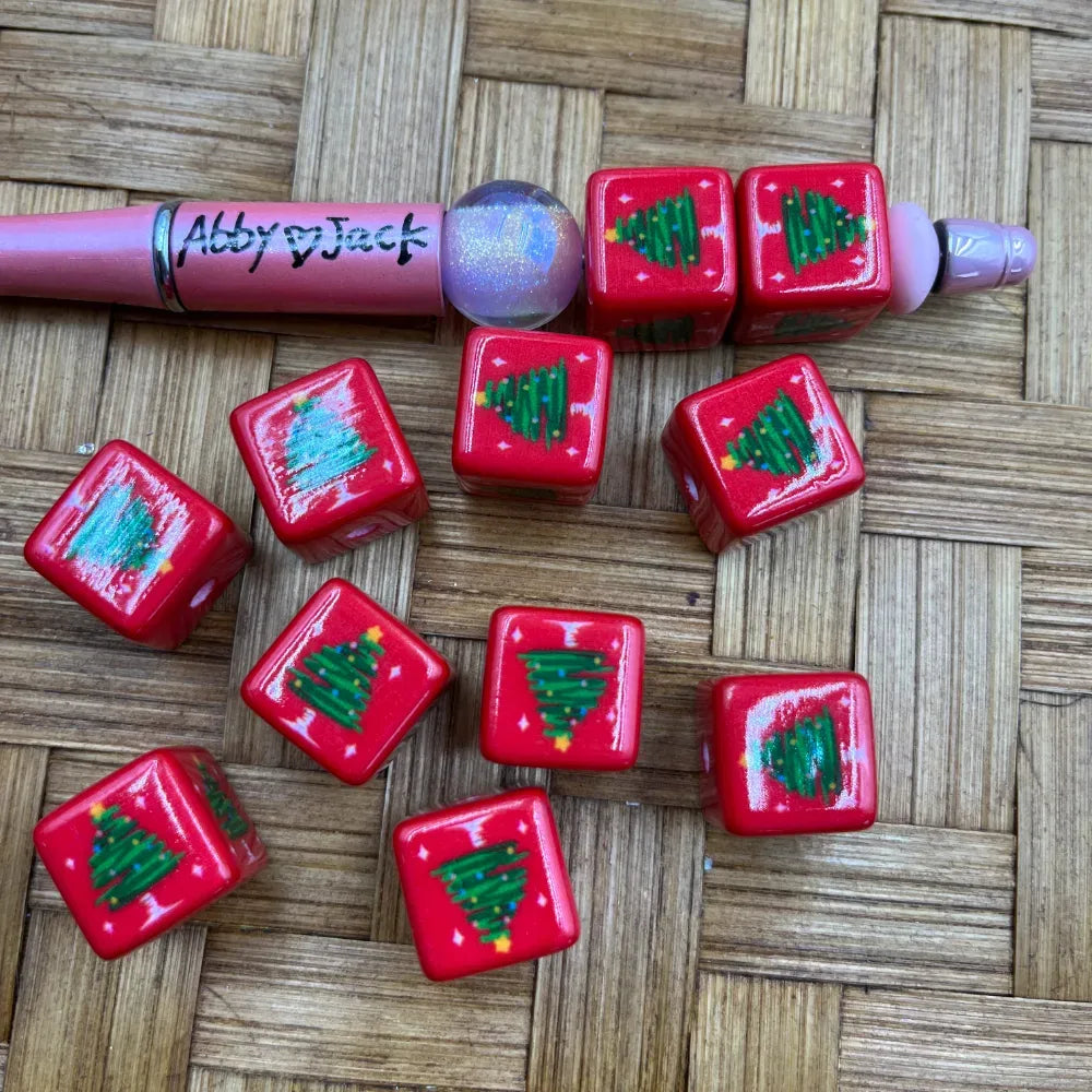 Red square acrylic beads featuring a green Christmas tree design with colorful lights, shown with a pink beadable pen on a woven bamboo surface.