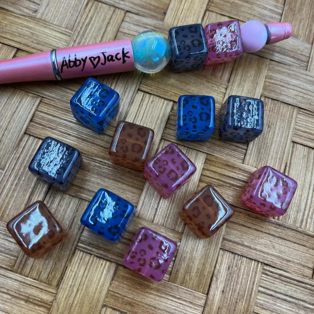 Leopard print square acrylic beads in blue, pink, brown, and black with metallic shimmer finish, displayed on a wicker surface next to a beadable pen.