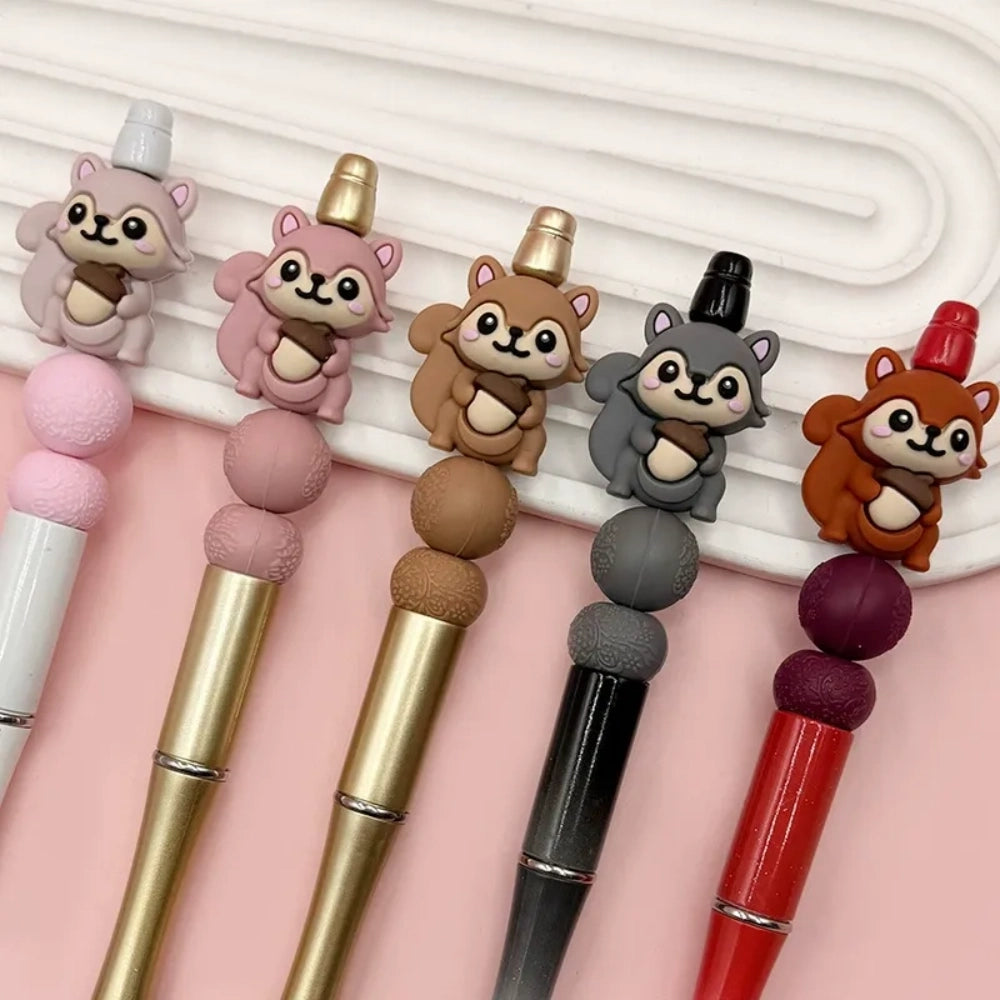 Five cute 3D squirrel silicone focal beads holding acorns in gray, pink, brown, dark gray, and red-brown colors displayed on decorative beadable pens against a pink background.