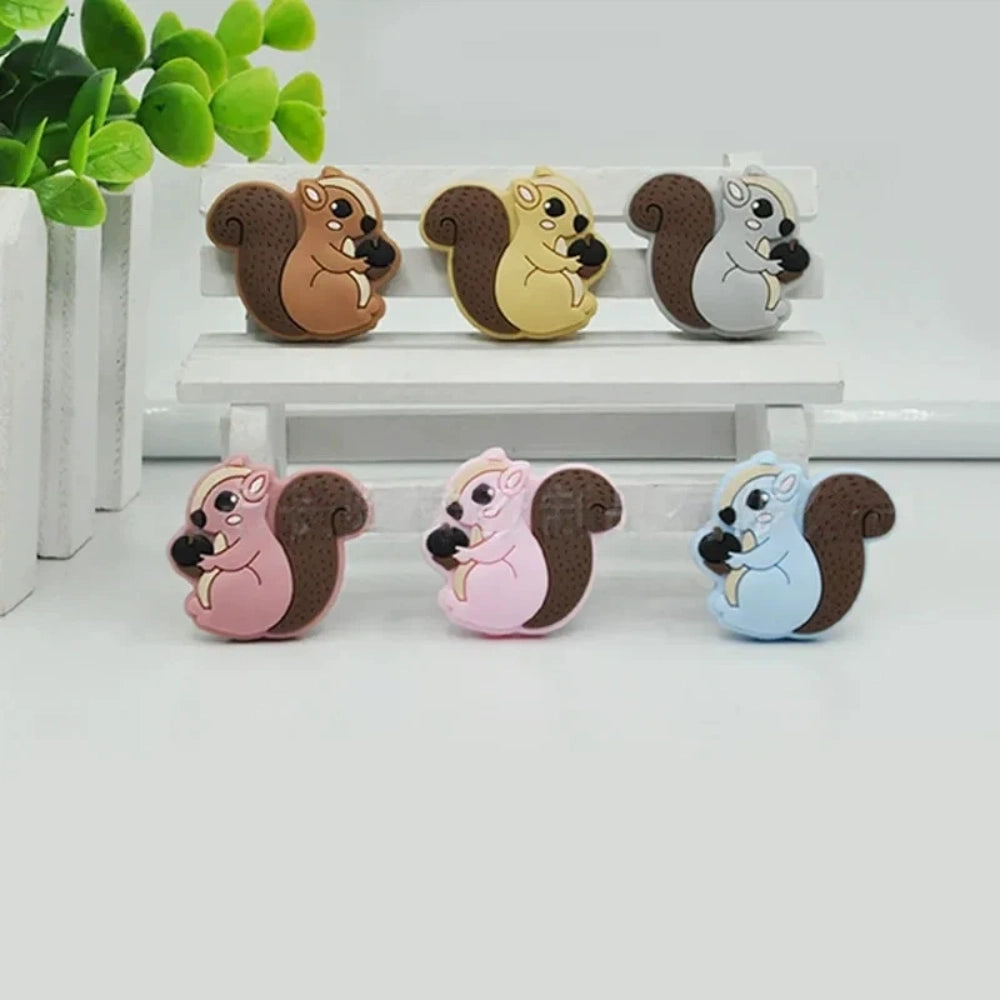 Six cute cartoon squirrel silicone focal beads holding acorns in brown, yellow, grey, dark pink, light pink, and blue colors arranged on a miniature white bench.