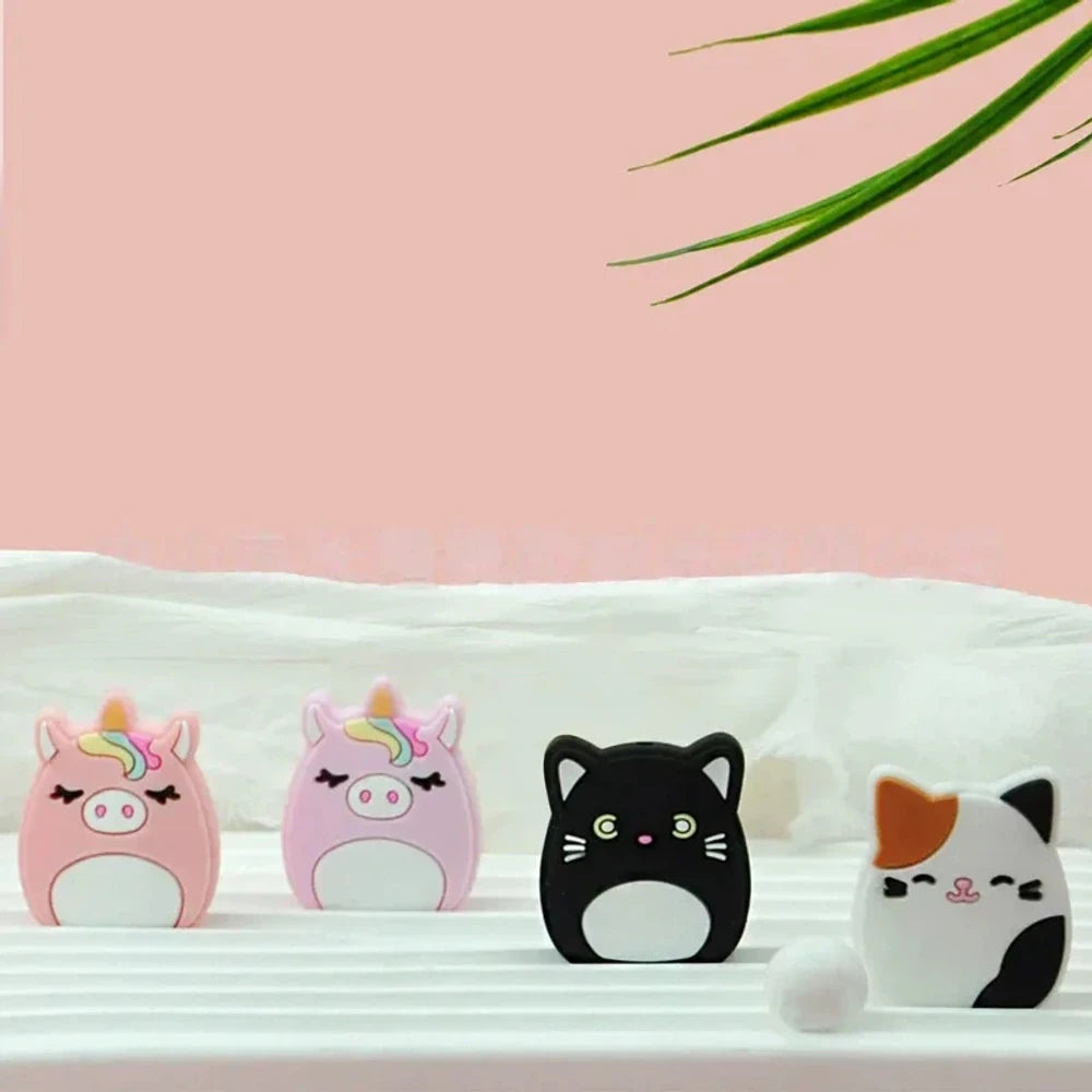 Four Squishmallow-style silicone focal beads featuring two pink unicorn pig characters and two cat characters (black and calico), displayed on a white surface with a pink background.