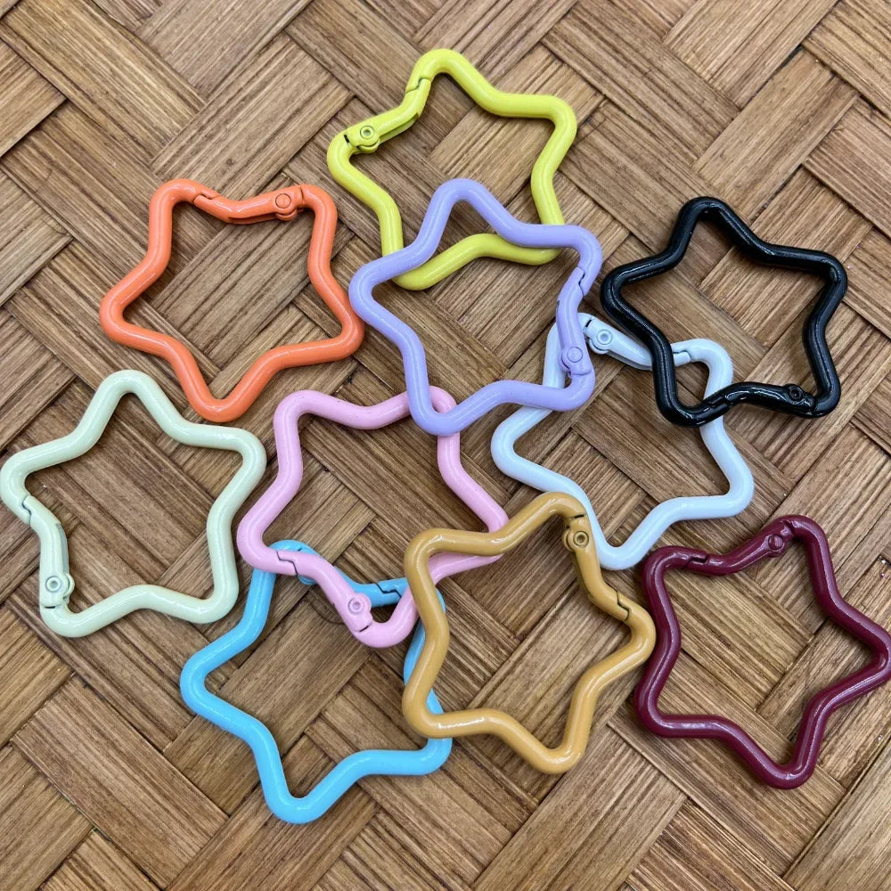 Colorful star-shaped carabiner clip keychain rings in orange, yellow, lavender, pink, blue, white, tan, black, maroon, and light green arranged on a woven surface.