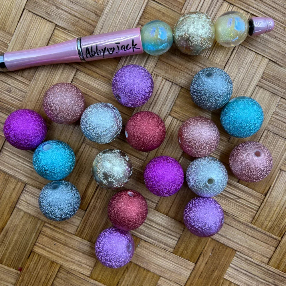 Assorted stardust metallic glitter acrylic beads in purple, teal, pink, rose gold, and gold colors displayed on a wicker surface next to a beadable pen labeled Abby Jack.