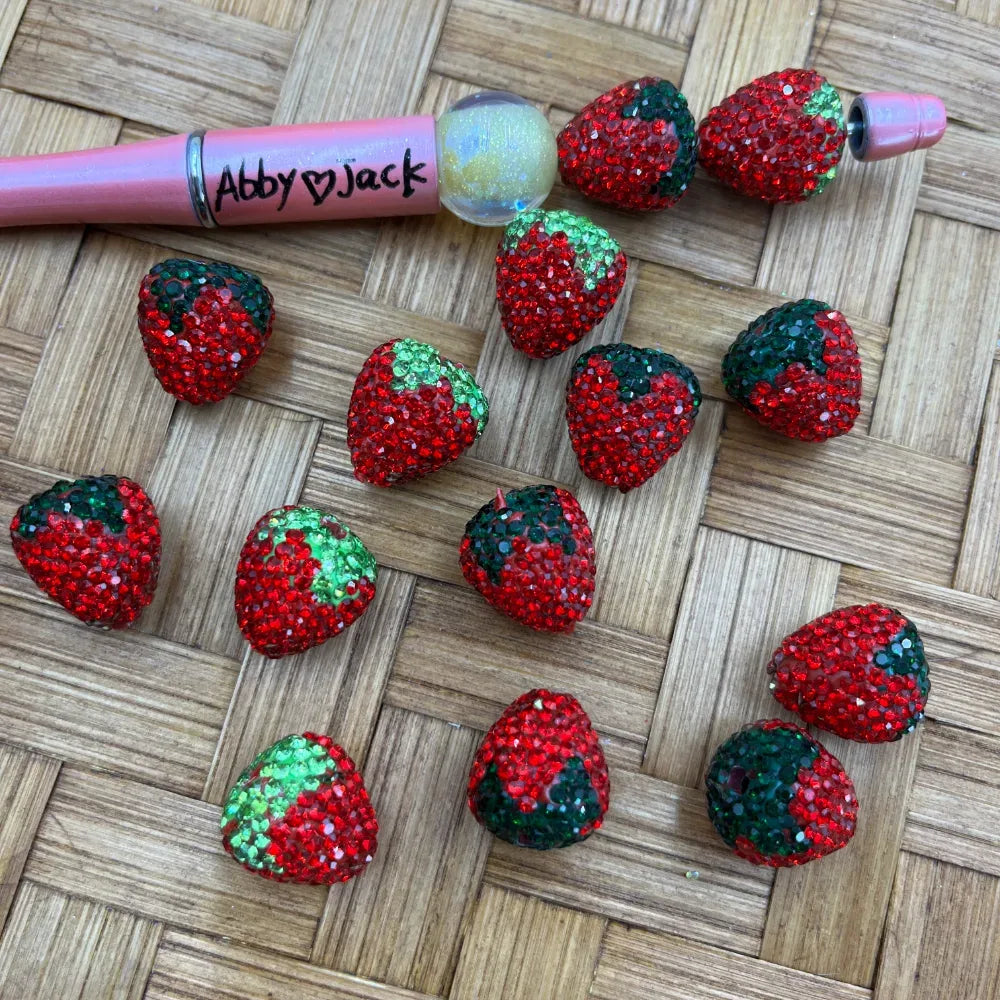 Sparkly strawberry-shaped clay rhinestone beads covered in red and green crystals displayed next to a pink beadable pen on a woven surface