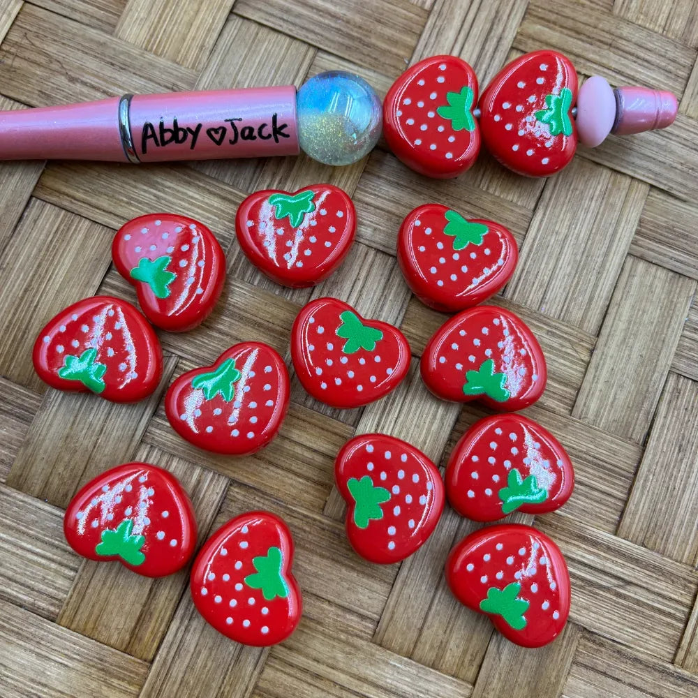 Red heart-shaped strawberry acrylic beads with green leaf tops and white polka dot details, displayed on a wicker surface next to a pink beadable pen labeled Abby and Jack.
