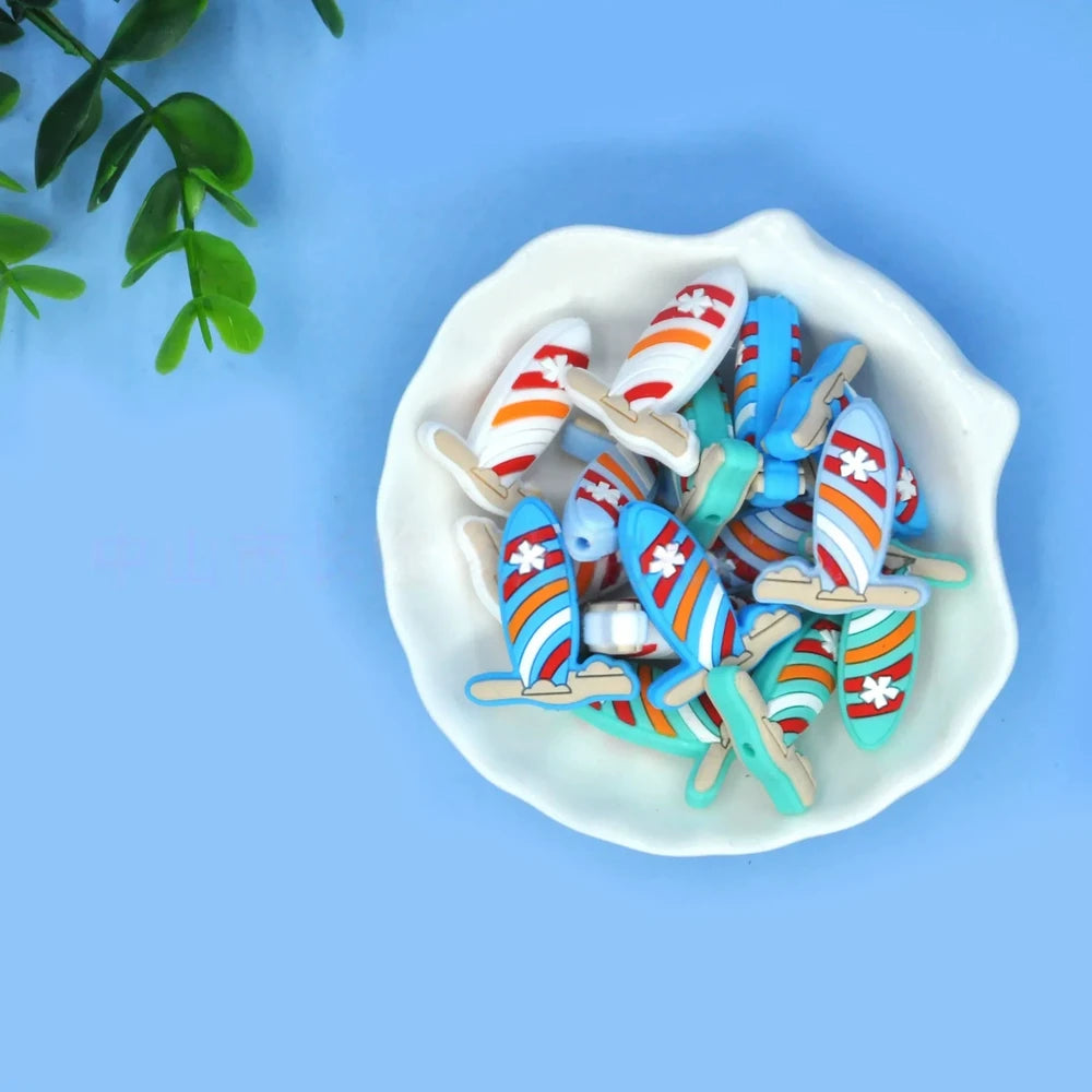 Colorful striped surfboard silicone focal beads in blue and white with flower detail, displayed in a white shell-shaped dish on a light blue background.
