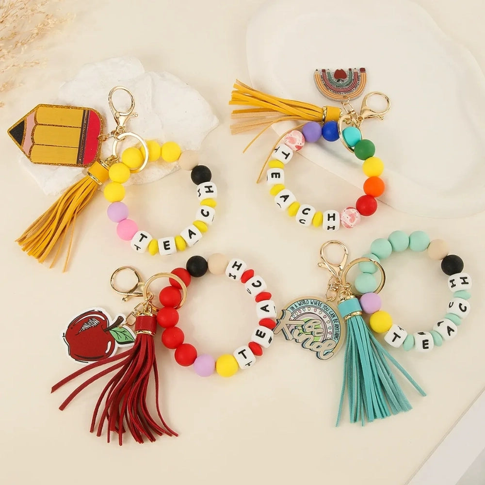 Colorful TEACH letter printed silicone beads on beaded bracelet keychains with tassels and teacher-themed charms including pencil, apple, rainbow, and Be Kind pendants.