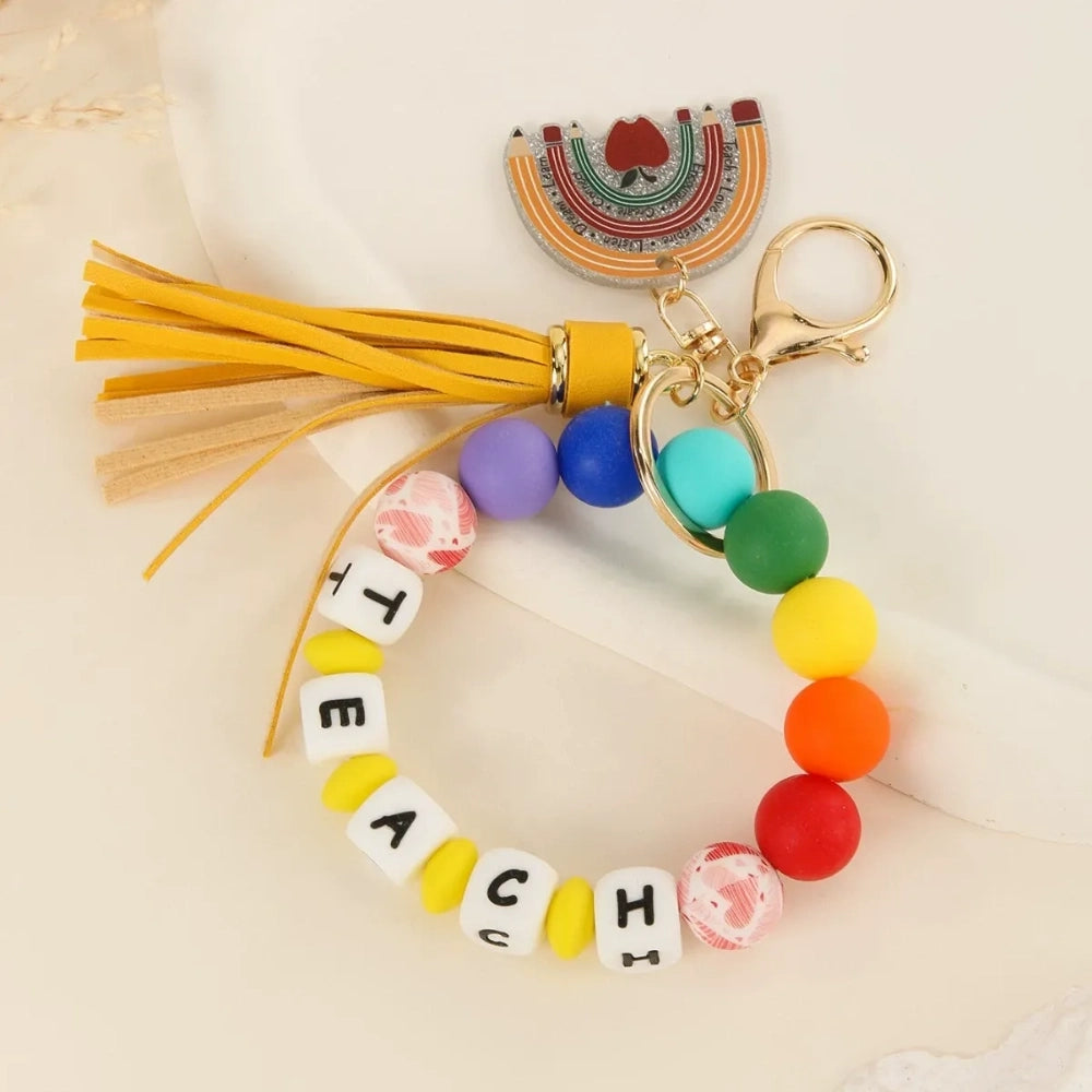 Colorful TEACH letter printed silicone beads on beaded bracelet keychains with tassels and teacher-themed charms including pencil, apple, rainbow, and Be Kind pendants. - A