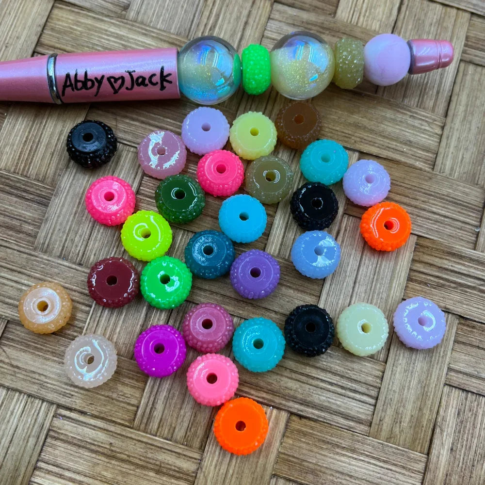 Assorted colorful textured rhinestone rim acrylic spacer beads in multiple colors including neon, pastel, and black, displayed on a woven surface next to a pink beadable pen labeled Abby Jack.