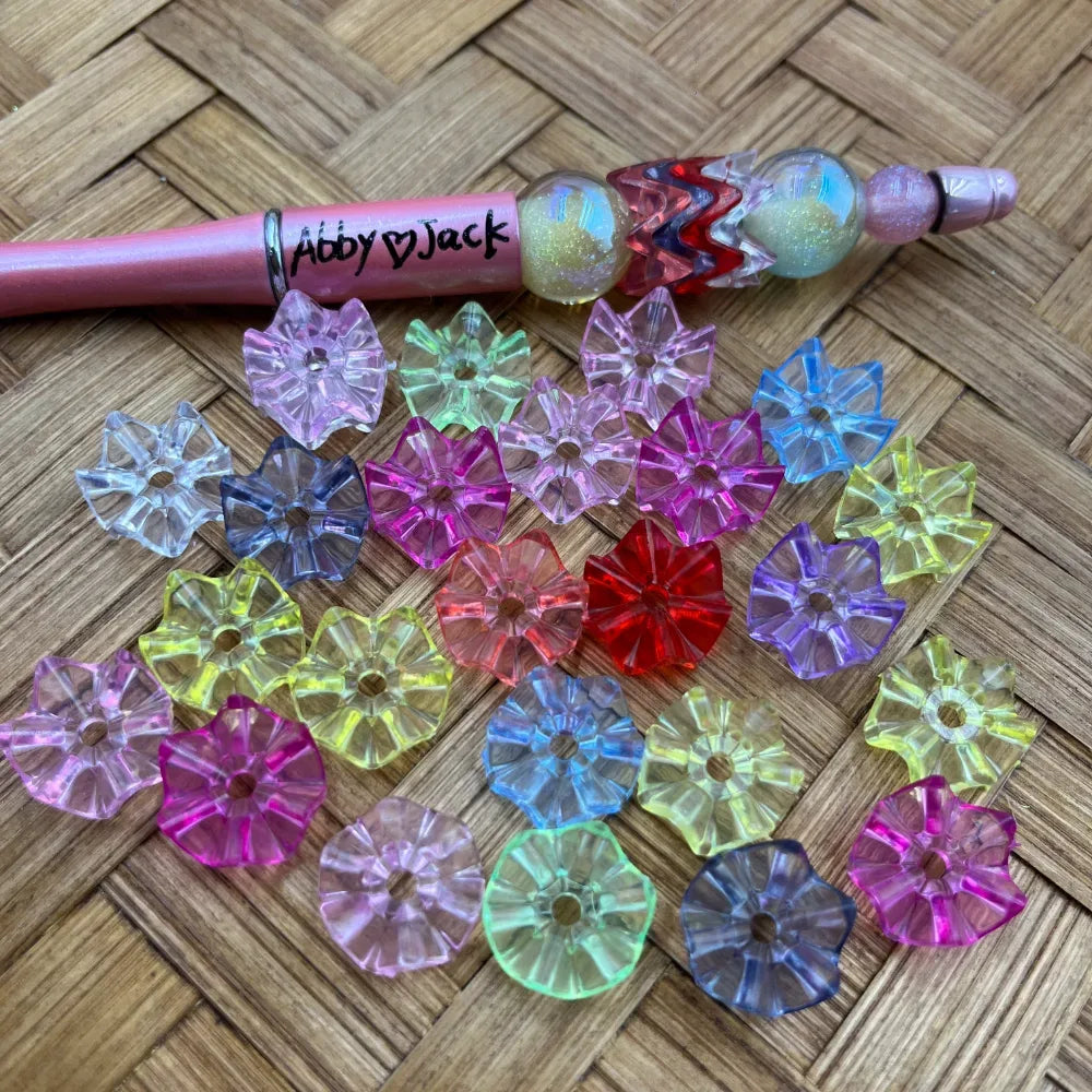 Assorted transparent flower-shaped acrylic spacer beads in multiple colors including pink, blue, yellow, red, purple, and green displayed on a woven surface next to a pink beadable pen labeled Abby and Jack.