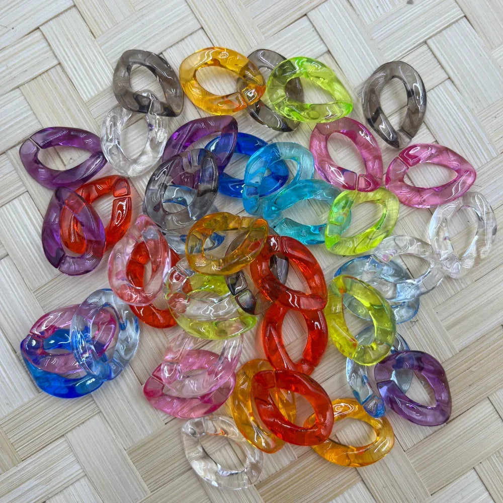Assorted colorful transparent oval chain link acrylic spacer beads in multiple colors including red, blue, pink, orange, purple, green, yellow, and clear, spread on a woven surface for DIY jewelry making.