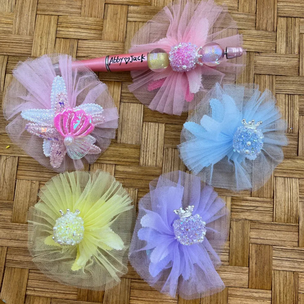 Pastel tulle pom flower fancy beads with crown and pearl centers in pink, blue, yellow, lavender, and mauve colors displayed on woven rattan surface with a decorated beadable pen