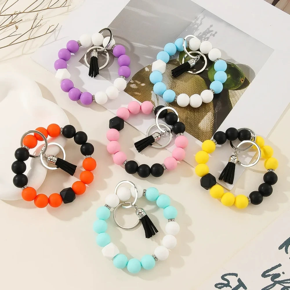 Six two-tone solid silicone bead keychain bracelets with black tassel and silver ring in purple-white, blue-white, pink-black, orange-black, yellow-black, and teal-white color combinations on a white surface.