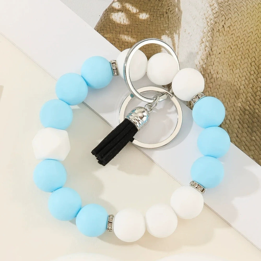 Six two-tone solid silicone bead keychain bracelets with black tassel and silver ring in purple-white, blue-white, pink-black, orange-black, yellow-black, and teal-white color combinations on a white surface. - A