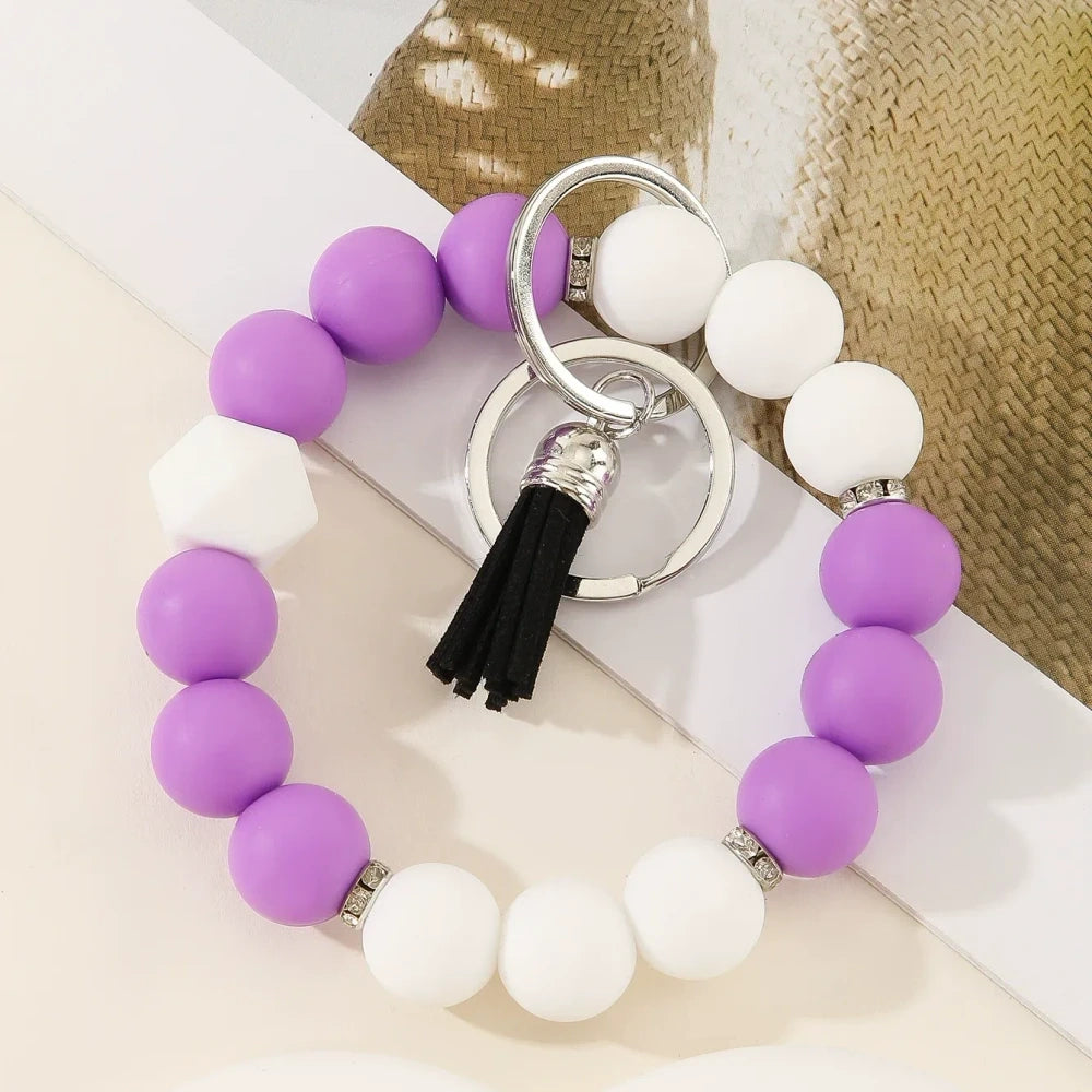 Six two-tone solid silicone bead keychain bracelets with black tassel and silver ring in purple-white, blue-white, pink-black, orange-black, yellow-black, and teal-white color combinations on a white surface. - B