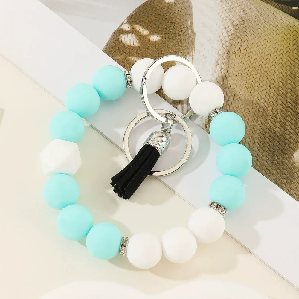 Six two-tone solid silicone bead keychain bracelets with black tassel and silver ring in purple-white, blue-white, pink-black, orange-black, yellow-black, and teal-white color combinations on a white surface. - C