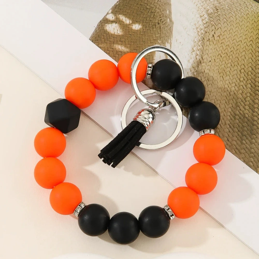 Six two-tone solid silicone bead keychain bracelets with black tassel and silver ring in purple-white, blue-white, pink-black, orange-black, yellow-black, and teal-white color combinations on a white surface. - D