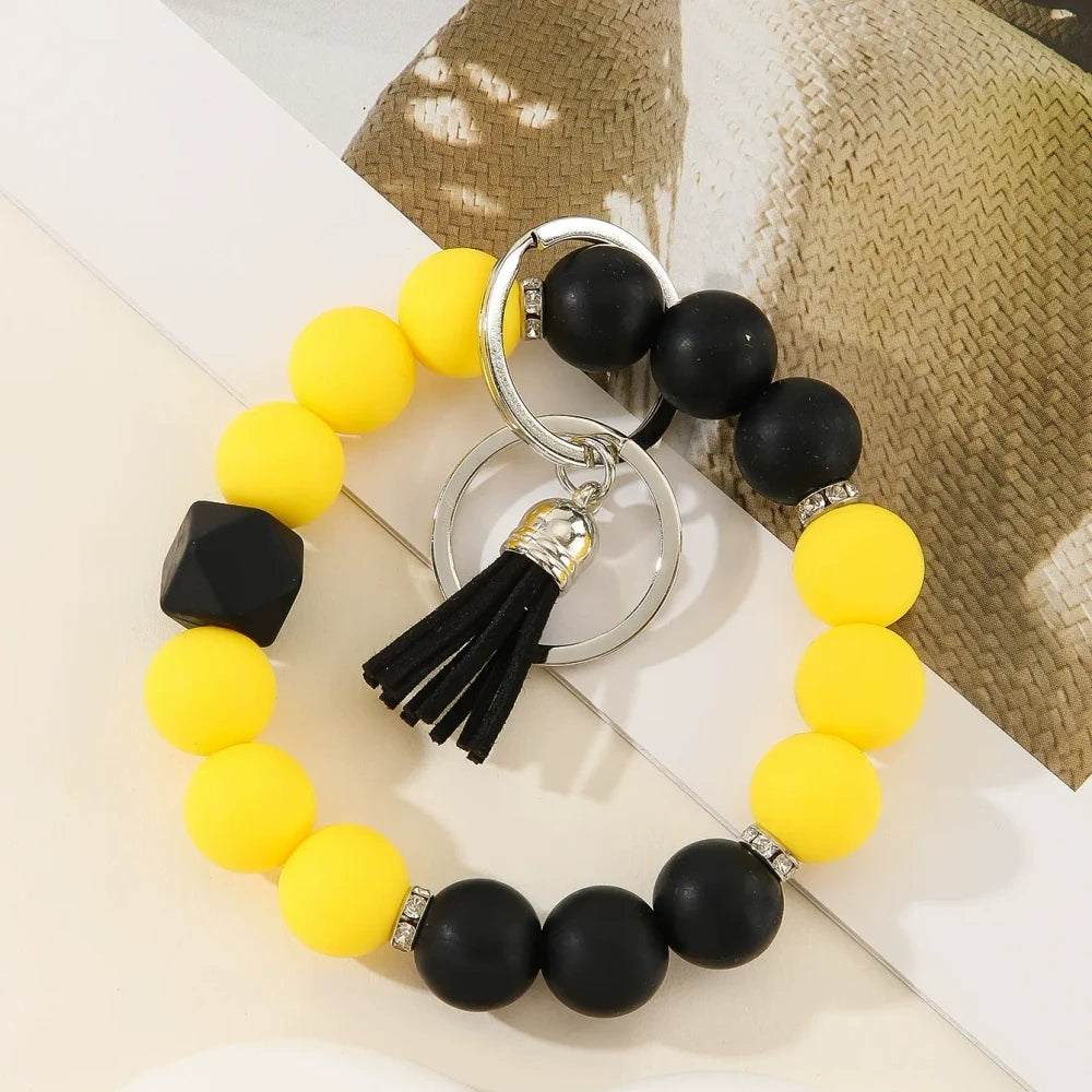 Six two-tone solid silicone bead keychain bracelets with black tassel and silver ring in purple-white, blue-white, pink-black, orange-black, yellow-black, and teal-white color combinations on a white surface. - E