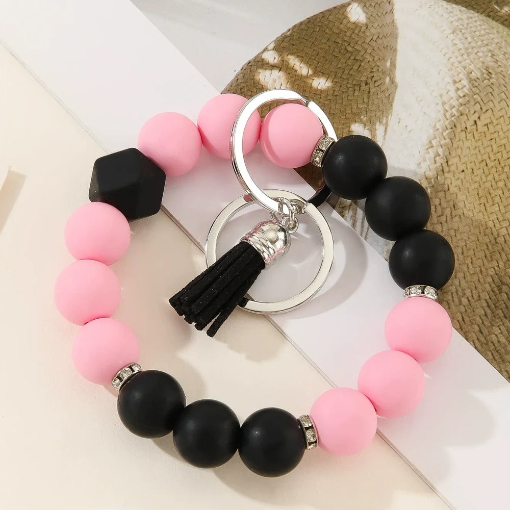 Six two-tone solid silicone bead keychain bracelets with black tassel and silver ring in purple-white, blue-white, pink-black, orange-black, yellow-black, and teal-white color combinations on a white surface. - F