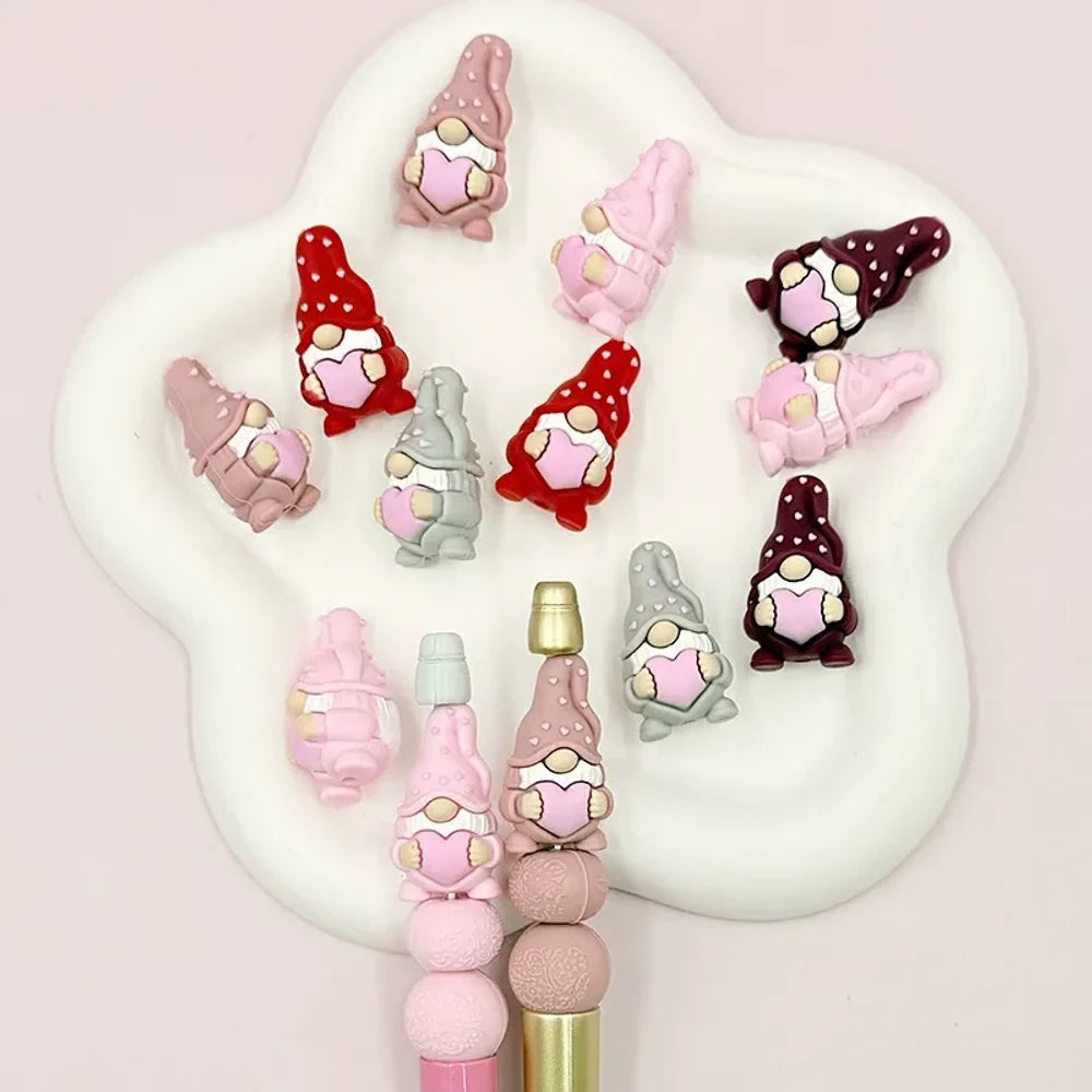 Assorted Valentine gnome silicone focal beads in red, pink, gray, and burgundy holding pink hearts, displayed on a white cloud tray with beadable pens below.