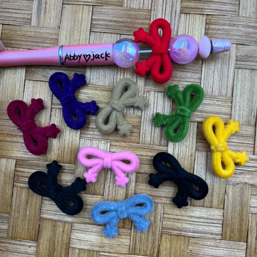 Assorted velvet flocked bow-shaped acrylic beads in multiple colors including red, pink, blue, black, green, yellow, navy, burgundy, tan, and dark teal displayed on a woven surface next to a pink beadable pen labeled Abby Jack.