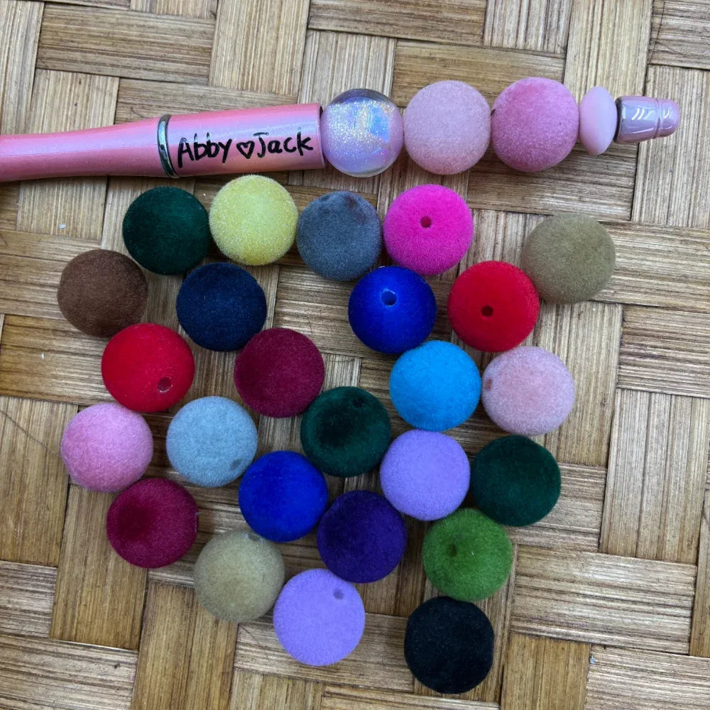 Assorted velvet flocked round acrylic beads in multiple colors including pink, blue, green, red, purple, and neutral tones displayed on a woven surface next to a beadable pen.