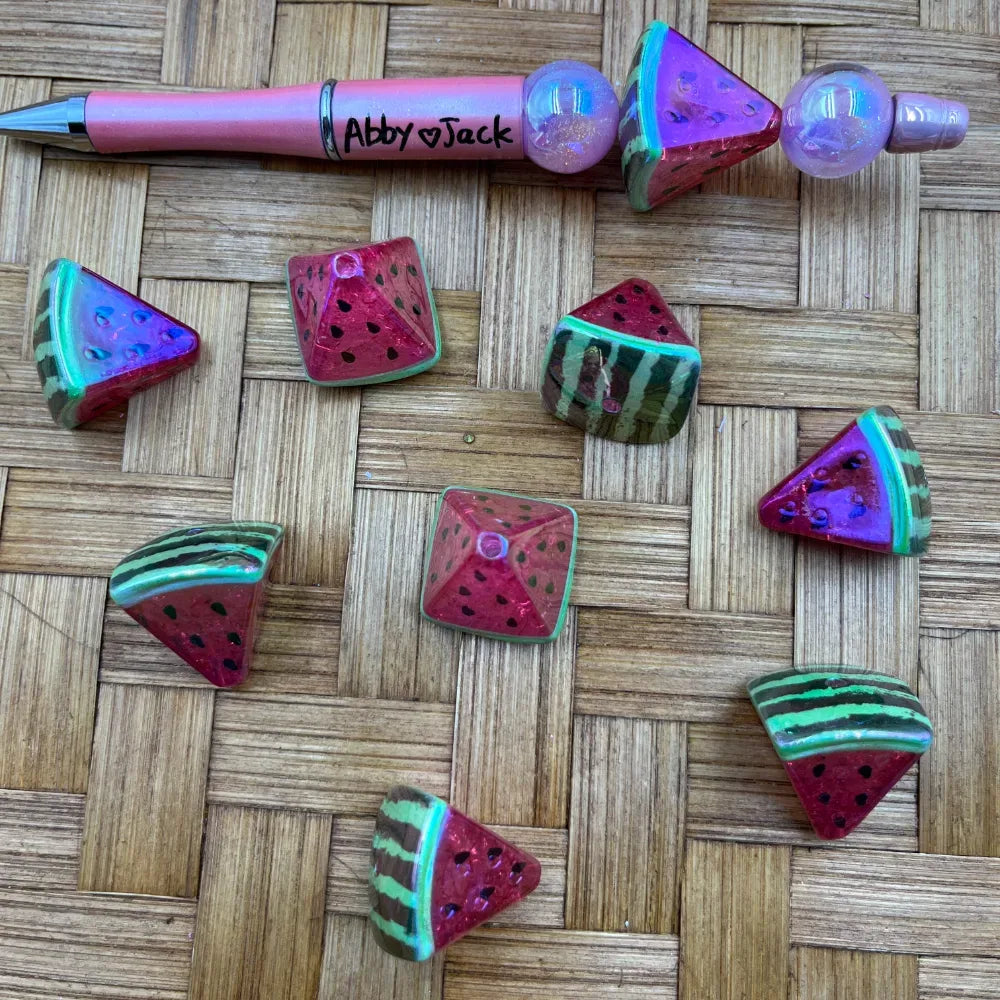 Watermelon slice shaped acrylic beads in red, green, and purple iridescent finish displayed next to a pink beadable pen on a woven bamboo surface.