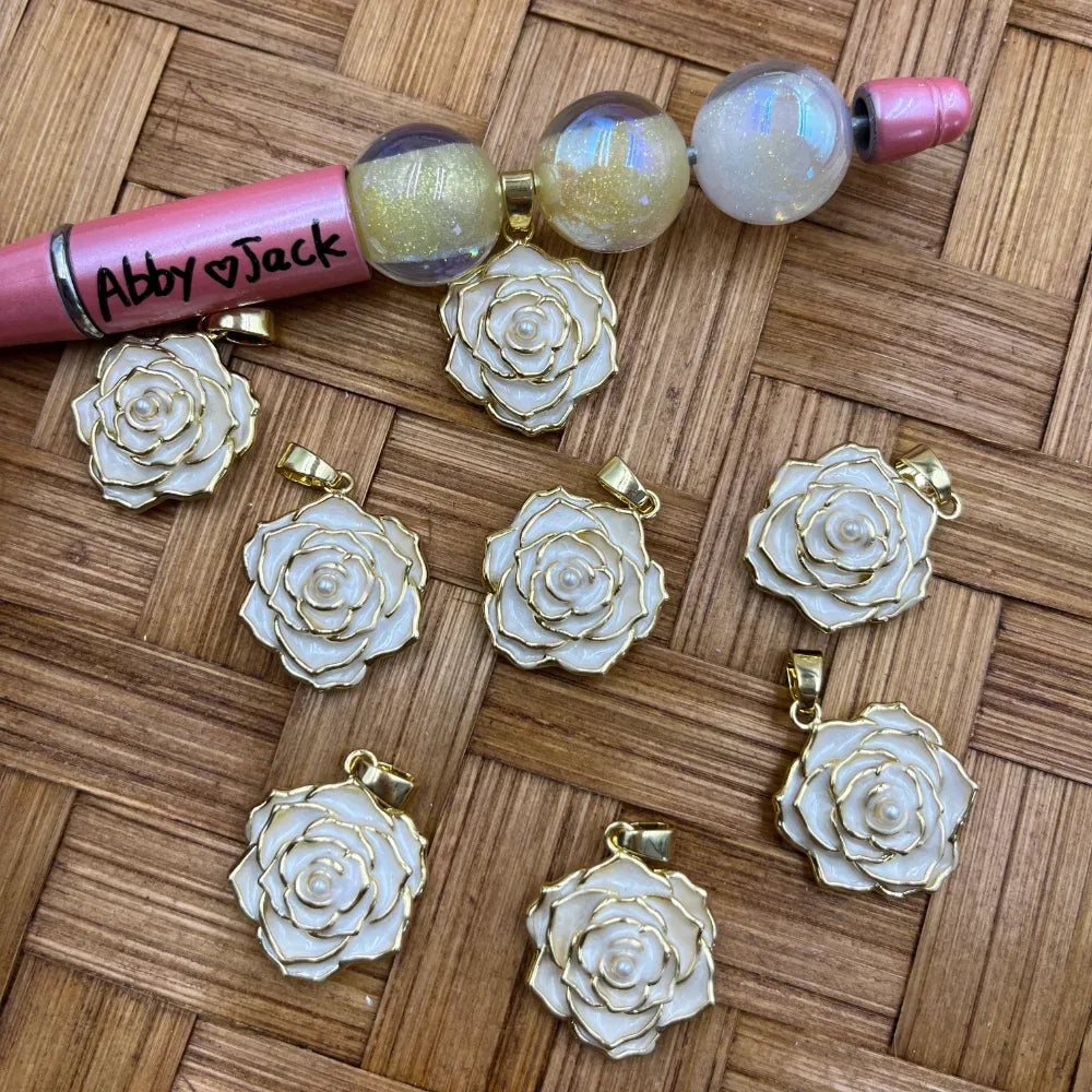White enamel rose flower bail pendants with gold trim and pearl center displayed on wicker surface next to a beadable pen, suitable for DIY jewelry making and beadable pen crafts.