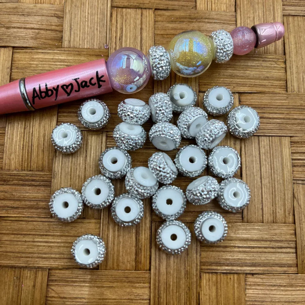 White acrylic spacer beads covered in silver rhinestone crystals with a center hole, shown with a beadable pen on a wicker surface