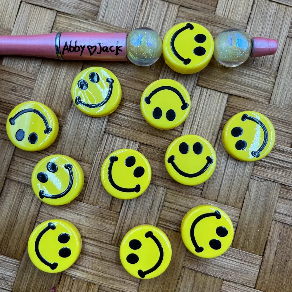 Yellow smiley face acrylic beads with black hand-drawn style happy face design arranged on a woven surface next to a beadable pen labeled Abby loves Jack and glitter beads.